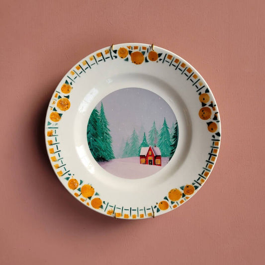 Een decoratief vintage wandbord met een illustratie van een rood winterhuisje en een oranje met groene rand.