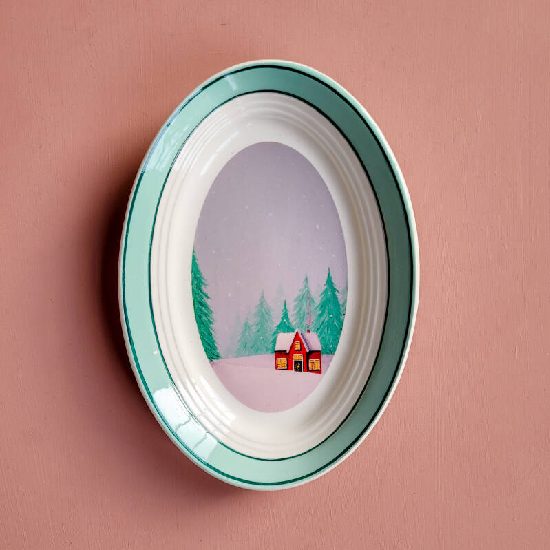 Een illustratie van een rood winterhuisje op een ovaal wandbord.