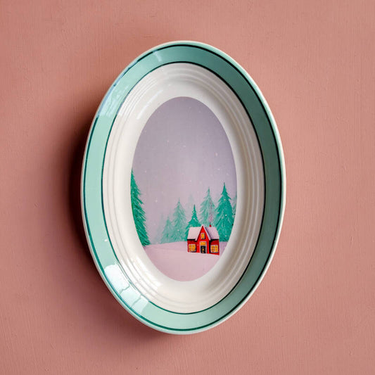 Een illustratie van een rood winterhuisje op een ovaal wandbord.